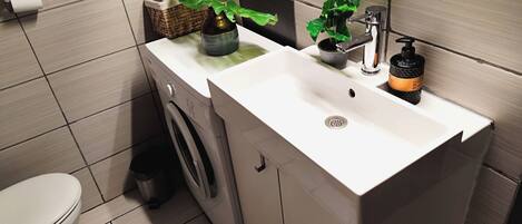 A bathroom with a white sink, a washing machine, a toilet, and a potted plant.