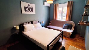 Quarto com cama, sofá, luminária, quadro de uma câmera e janela com cortinas.