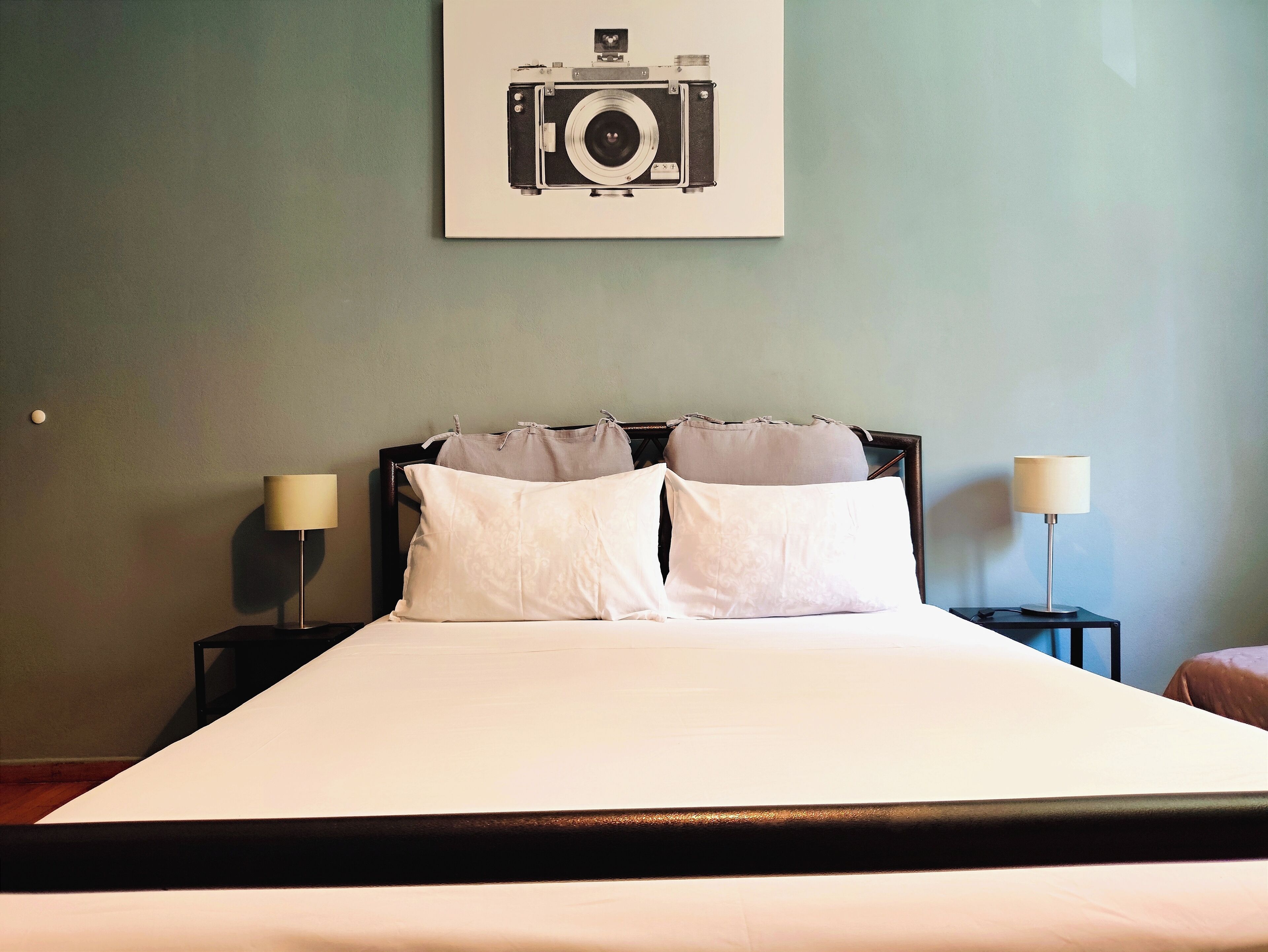 A neatly made bed with white linens, two bedside lamps, and a framed picture of a camera on the wall.