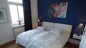 A bedroom with a large bed, a window with curtains, a radiator, a painting on the wall, and bedside lamps. - Morizan (Röbel/Müritz)
