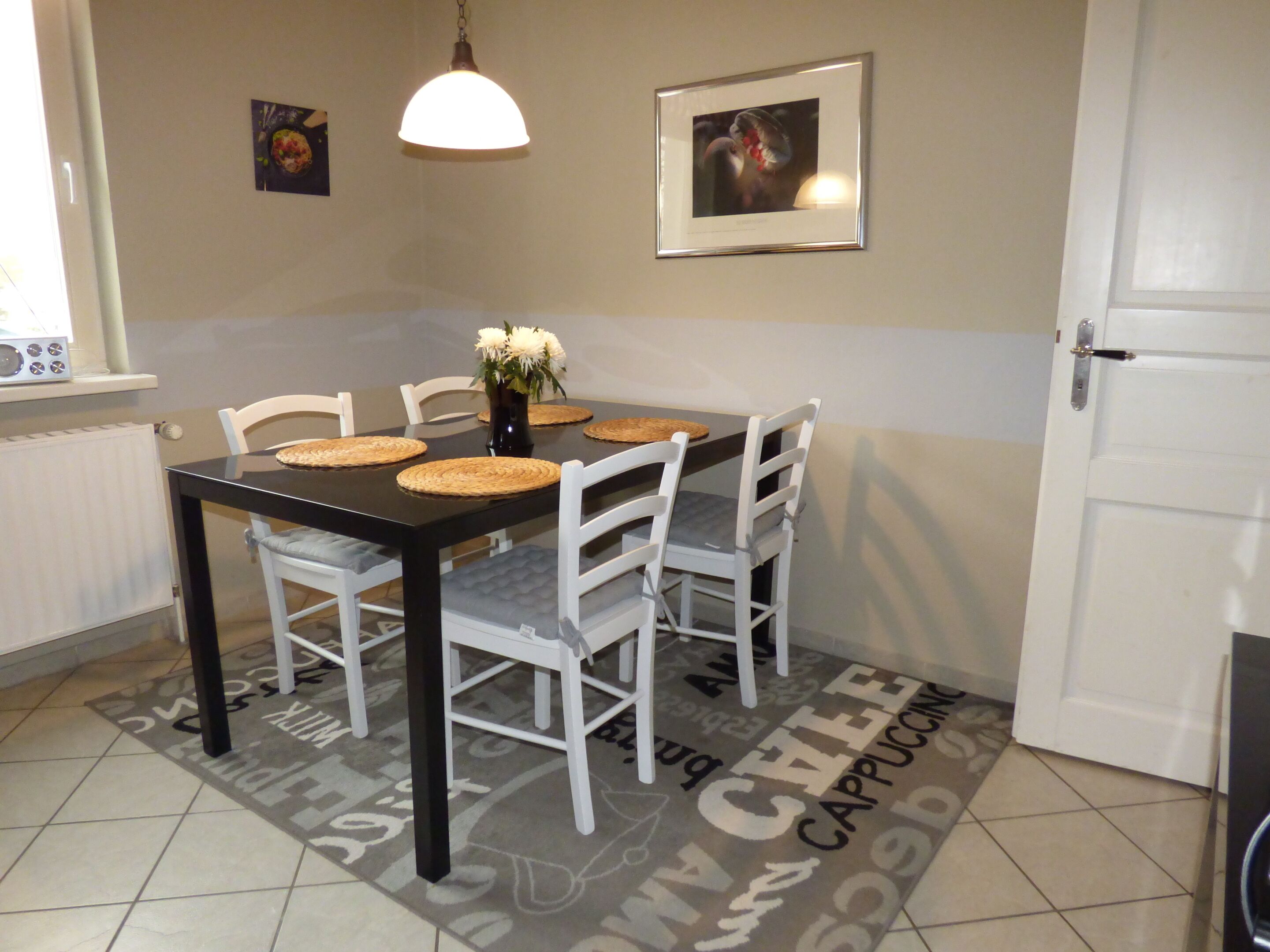 A dining area with a black table, white chairs, a vase with flowers, a framed picture on the wall, and a door to the right.