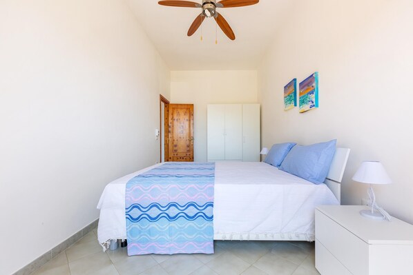Une chambre à coucher comprenant un lit, une table de chevet, un ventilateur de plafond, une porte en bois et deux œuvres d’art mural.