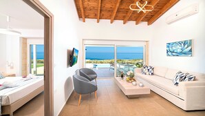 A modern room with a bed, a grey armchair, a sofa, a TV, and a large window with a view of the sea. - Vista Mare Maxima Heated Pool (Georgioupoli)