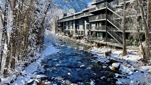 A modern building with multiple balconies overlooking a snow-covered river, surrounded by trees and mountains.