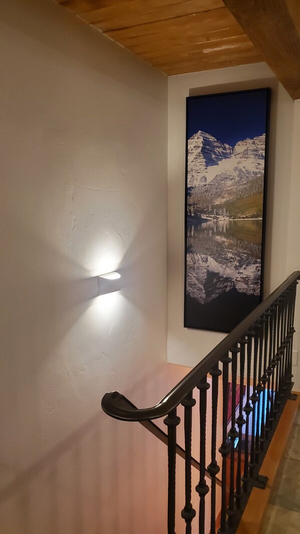 A staircase with a handrail, a wall-mounted light, and a framed picture of a mountain landscape. - Downtown Aspen Riverfront 3BR Luxury Duplex, Pool, Hot Tub, Gym, Walk to Gondola (Aspen)
