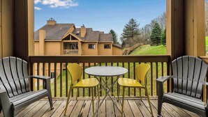 A wooden deck with two Adirondack chairs and a small round table, overlooking a house with a sloped roof and a chimney. - SnowPine Condo, Sleeps 6, King Bed, Patio (Ellicottville)
