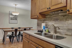 A kitchen with wooden cabinets, a granite countertop, a stainless steel sink, and a dining area with a table and chairs. - SnowPine Condo, Sleeps 6, King Bed, Patio (Ellicottville)