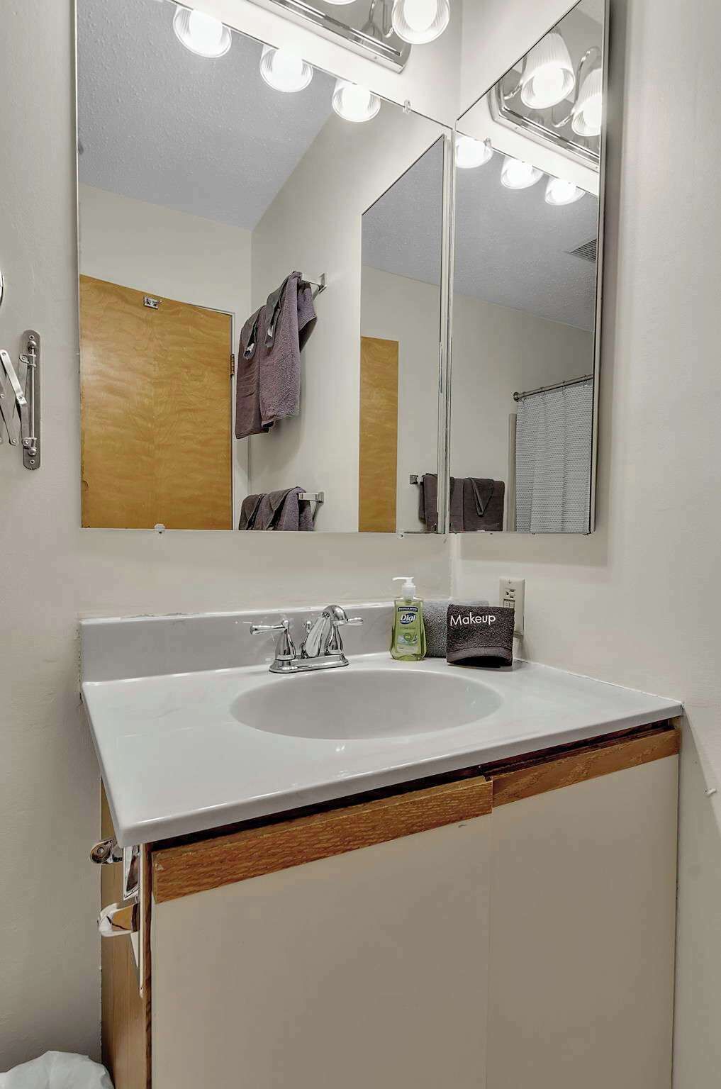 A bathroom with a white sink, a large mirror with lights, a wooden vanity, and a towel rack.