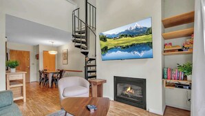A living room with a fireplace, a TV mounted on the wall, a staircase, and a dining area. - SnowPine Condo, Sleeps 6, King Bed, Patio (Ellicottville)