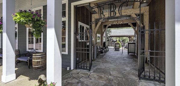 A covered outdoor area with a wooden bench, a hanging flower basket, and a stone pathway leading to a gate with 'BC' above it.