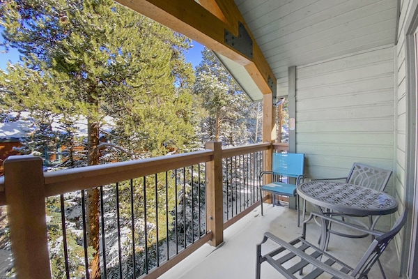 A wooden balcony with two metal chairs, a round table, and a blue box overlooks a forested area. The balcony has a roof structure supported by wooden beams. Snow is visible on the trees.