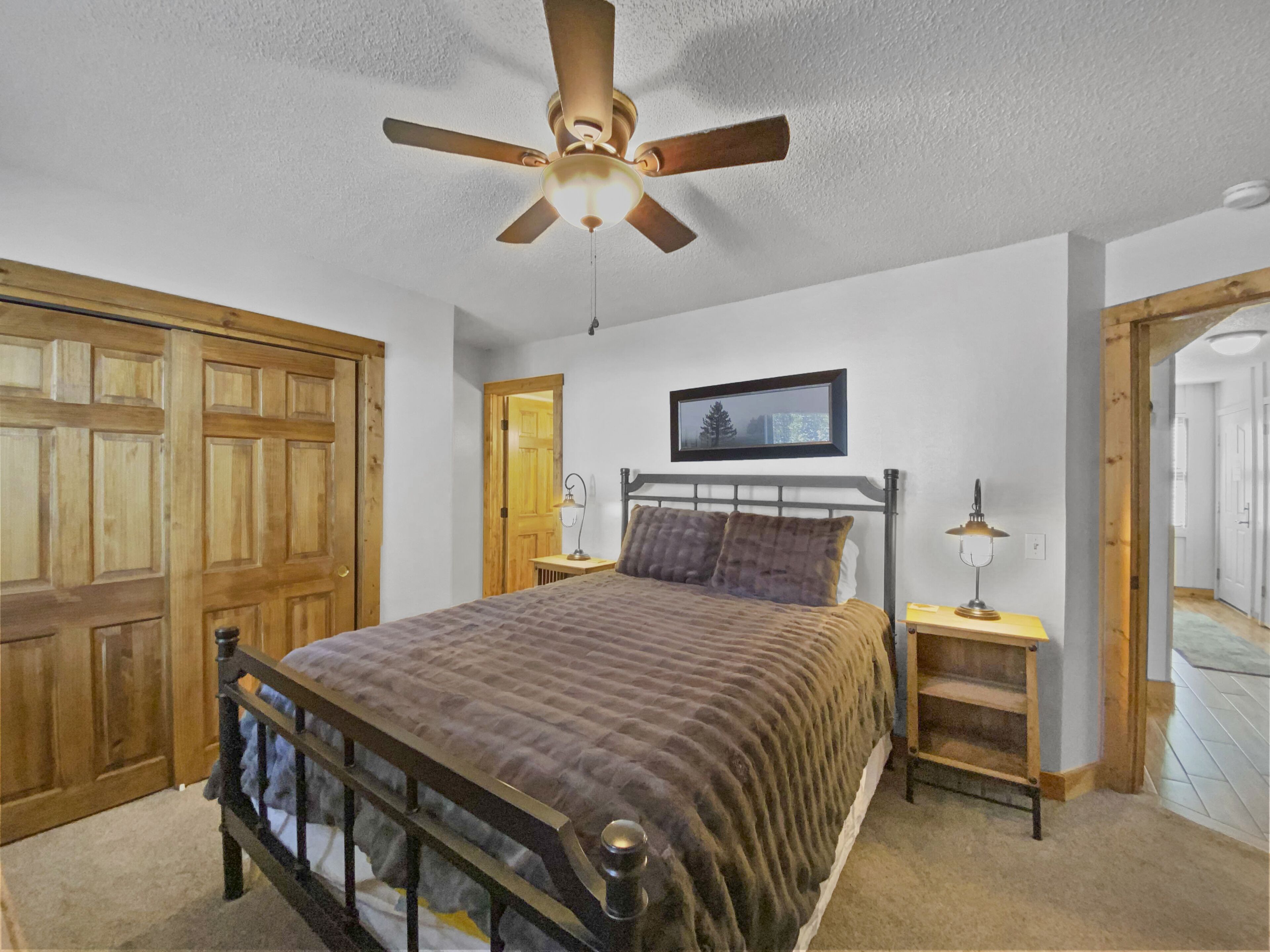 A bedroom with a bed, wooden wardrobe, ceiling fan, bedside table, and a framed picture on the wall.