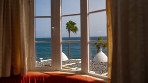 Een kamer met een groot raam, uitzicht op een kiezelstrand en een palmboom.
