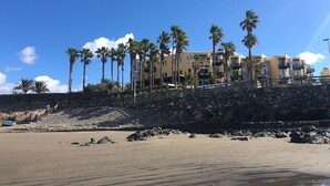 Hotel aan het strand met een stenen steunmuur, palmbomen en een helderblauwe lucht.