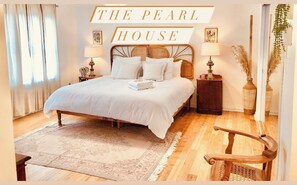 A neatly arranged bedroom with a large bed, bedside lamps, a decorative mirror, and a sign reading "THE PEARL HOUSE".