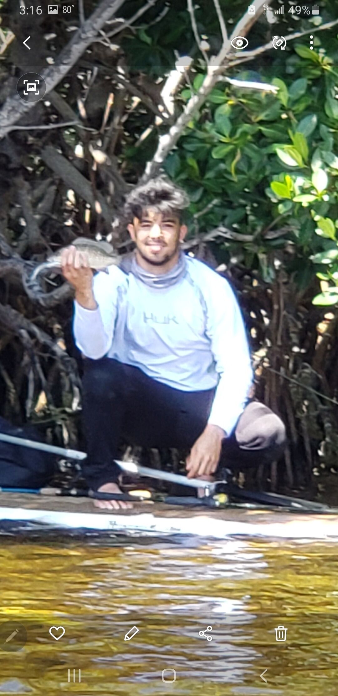 Una persona sosteniendo un pez cerca de un cuerpo de agua rodeado de vegetación densa.