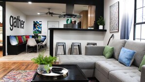 A modern living room with a sectional sofa, a coffee table with remote controls, a colorful rug, and a wall with the word "gather". - *Get-Away by River Trail, Hot Tub, Fire Pit, Bikes, Grill (Kerrville)