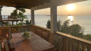 Een houten terras buiten met een tafel en stoelen, uitkijkend op een rustige zee en een zonsondergang.