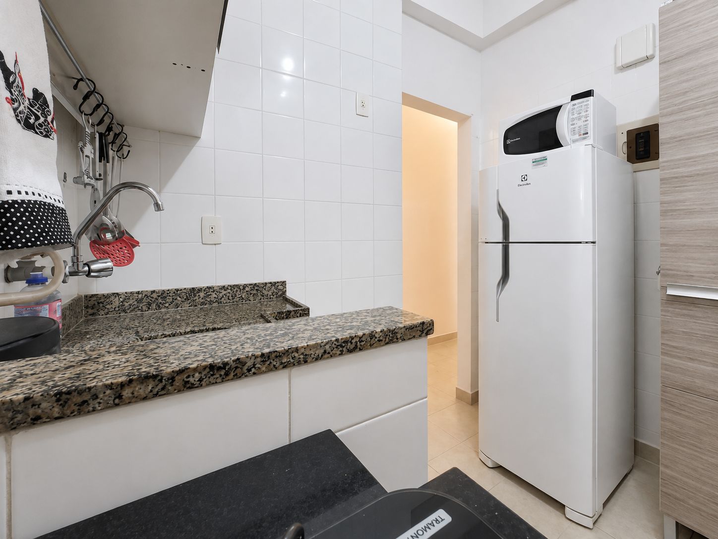 A kitchen with white tiled walls, a granite countertop, a white refrigerator, and a sink with a faucet.