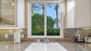 A kitchen with a granite countertop, a double sink, and a window with a view of palm trees. - Casa La Paz Getaway – Private Spa & BBQ,  Pool 2br (La Quinta)
