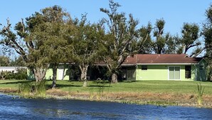 A house with a green exterior and a brown roof, surrounded by trees and a body of water.
