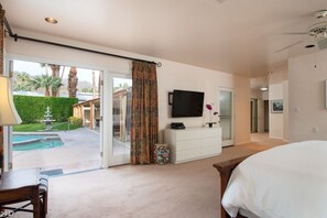 A hotel room with a large bed, a television, a desk, and a view of a swimming pool and garden. - Maison du Soleil (Palm Springs)