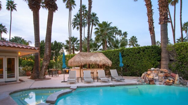 A swimming pool with lounge chairs, a thatched-roof structure, palm trees, and a rocky waterfall feature. - Maison du Soleil (Palm Springs)