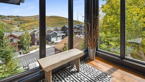 A room with a large window offering a view of a residential area and hills. A wooden bench and a potted plant are visible in the foreground. - Empire Villa (Park City)