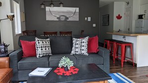 A modern living room with a grey sofa, red and patterned cushions, a coffee table with a plant and red decorations, a dining area with red stools, and a kitchen with a microwave and refrigerator.