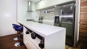 A modern kitchen with white cabinetry, a stainless steel refrigerator, and a bar stool with a blue seat. - ★18th Floor★Coolest building in Medellin★RoofTop Pool/Jacuzzi/Gym★Amazing Views★ (Medellin)
