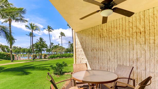 An outdoor patio with a round table and chairs, a ceiling fan, and a view of a well-maintained lawn and palm trees.