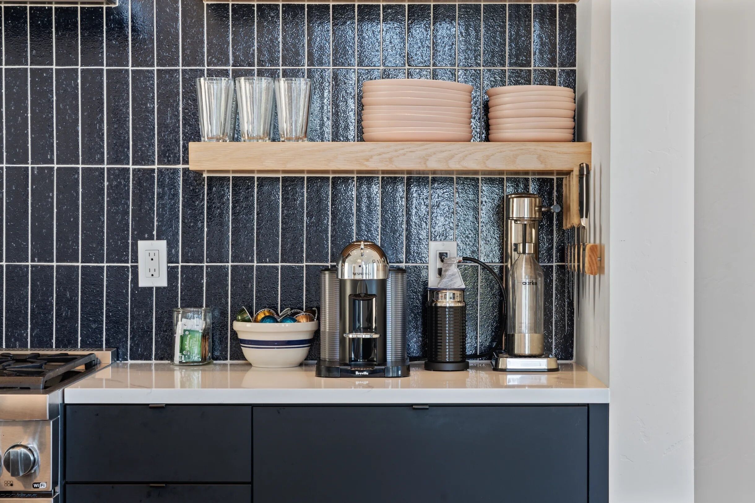 Une cuisine avec un plan de travail noir recouvert de carreaux, une étagère en bois où sont posées des assiettes et des verres, une machine à café, une bouilloire et une cuisinière.