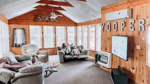A cozy log cabin interior with a ceiling fan, large windows, a TV, and a 'YOOPER' sign. - Lake House 100 yards off snowmobile trail #8.  (Michigamme)