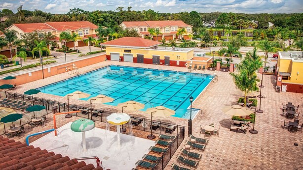 Una gran piscina con múltiples carriles, rodeada de sillones de descanso y sombrillas, con edificios residenciales al fondo.