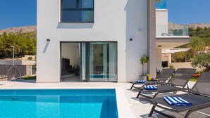 A modern pool area with a clear blue pool, lounge chairs with towels, and a building with large windows and a sliding door. - Villa Calypso with Pool (Podstrana)