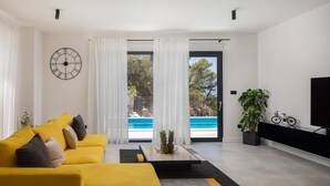 A modern living room with a yellow sofa, a coffee table with pineapples, a large clock, and a view of a swimming pool. - Villa Calypso with Pool (Podstrana)