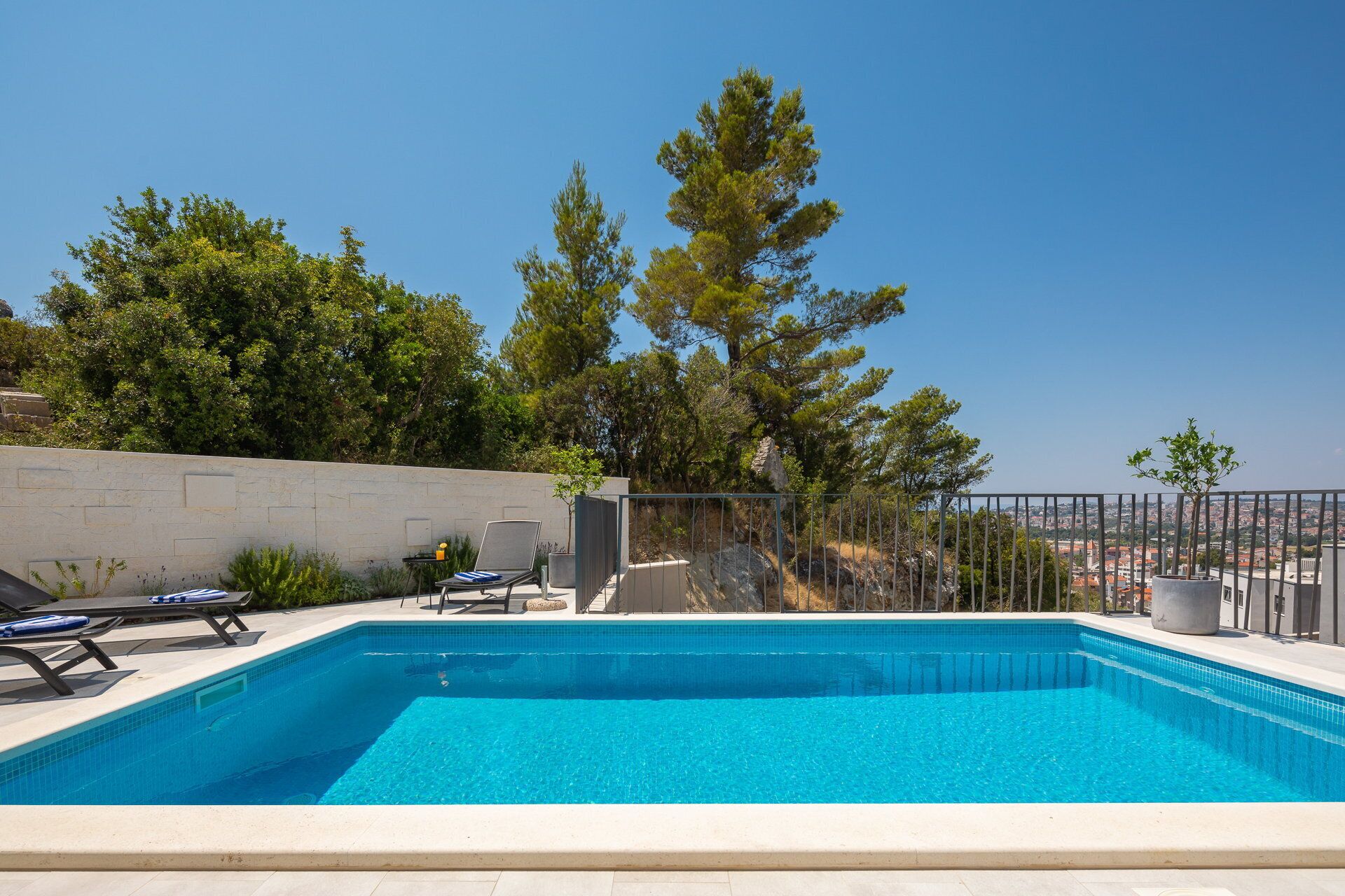 A rectangular swimming pool with clear blue water, surrounded by a low stone wall and a metal fence. There are lounge chairs and a small table with a yellow cup on the poolside.