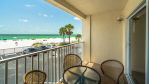 A balcony with wicker chairs and a table, overlooking a beach with palm trees and parked cars.