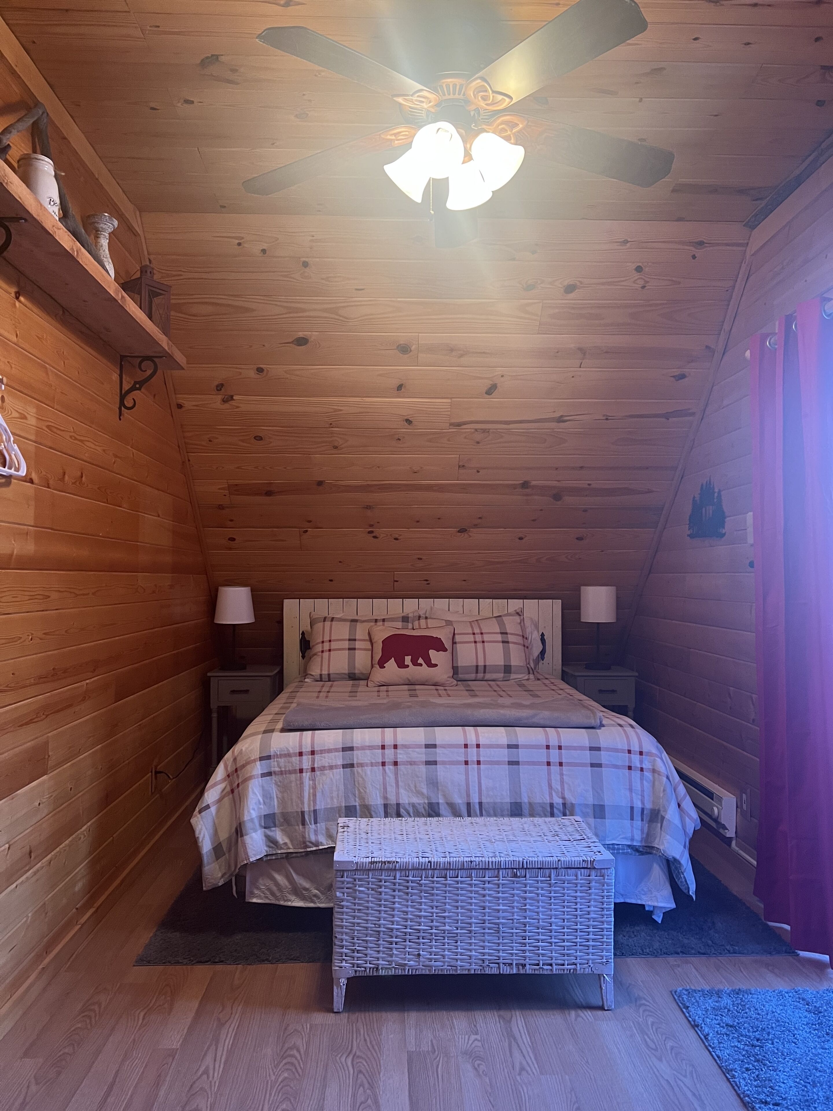 A cozy wooden cabin bedroom with a bed, two bedside tables, a ceiling fan, and a small bench.