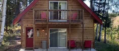 A wooden cabin with a red door, a balcony with red chairs, and a gravel driveway.