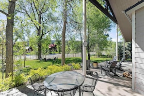 Une terrasse couverte avec une table et des chaises, un barbecue et une vue sur une cour verdoyante avec des arbres.