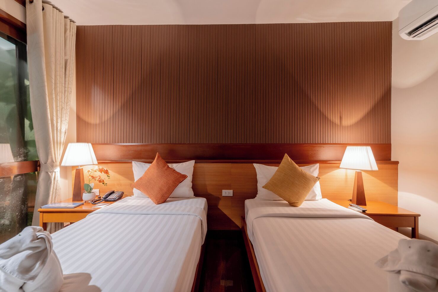 A hotel room with two beds, wooden headboards, bedside lamps, and a window with curtains.