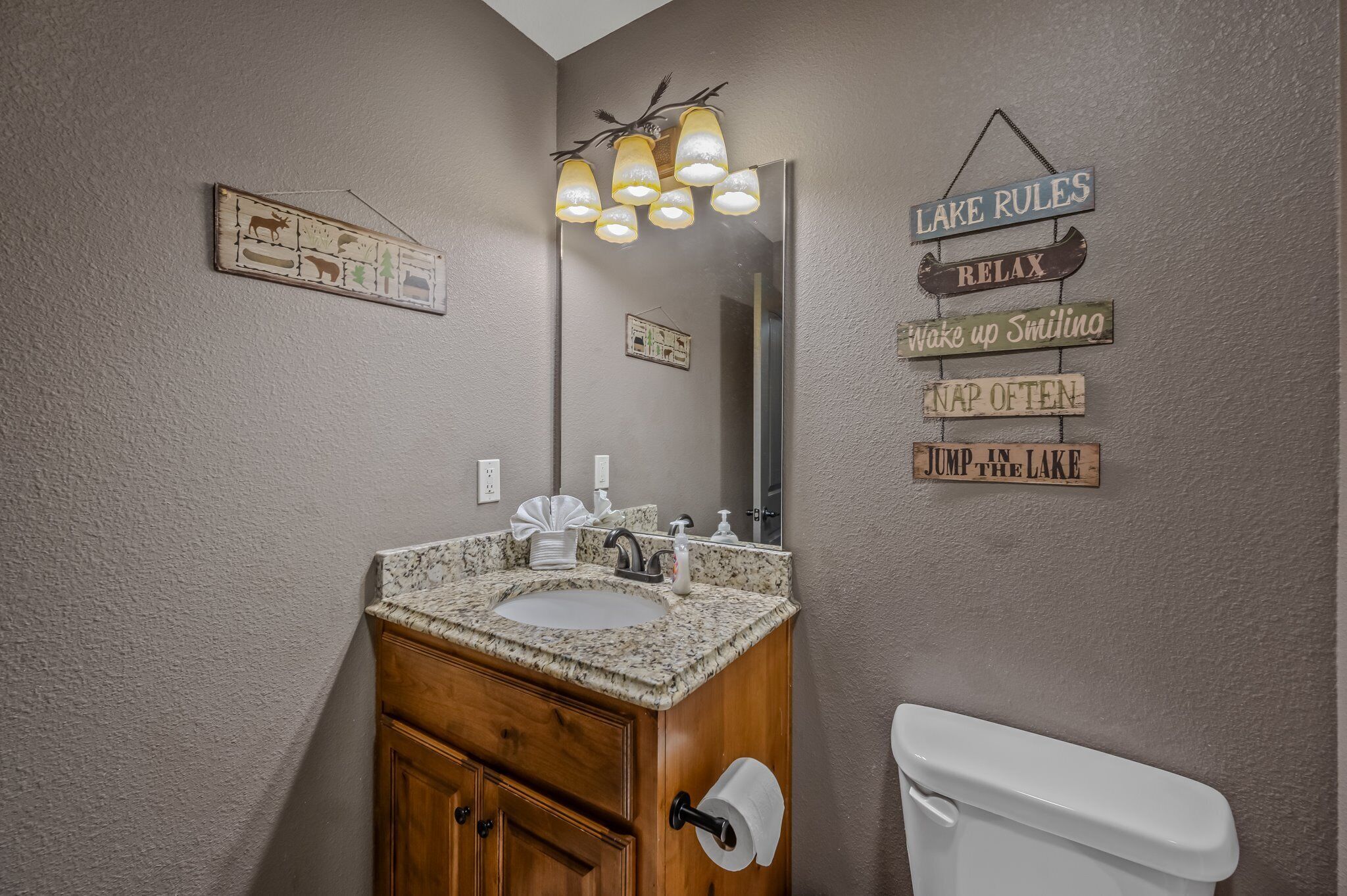 A bathroom with a granite sink, wooden cabinet, and a toilet. There are wall-mounted signs with phrases like "LAKE RULES" and "RELAX".