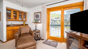 A living room with a brown leather sofa, wooden cabinets, a television, and a balcony with a view of greenery. - Family Friendly Condo- Walk to Main Street and Town Lift (Park City)