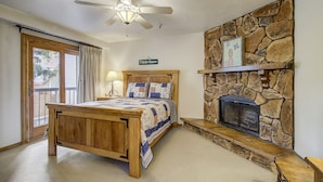 A bedroom with a wooden bed, a stone fireplace, a ceiling fan, and a large window with curtains. - Family Friendly Condo- Walk to Main Street and Town Lift (Park City)