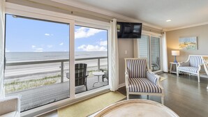 Ein Zimmer mit großem Fenster mit Blick auf einen Strand, einem an der Wand befestigten Fernseher und gestreiften Stühlen.