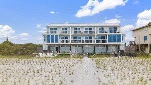 Ein mehrstöckiges Strandhaus mit Balkon, ein Sandstrand davor und ein klarer Himmel.
