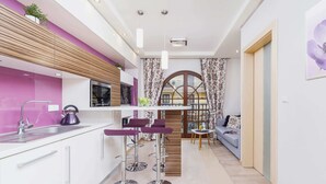 Une cuisine moderne avec un plan de travail violet, des armoires blanches et une bouilloire en acier inoxydable. On y trouve des tabourets de bar violets et une étagère en bois ornée de plantes.