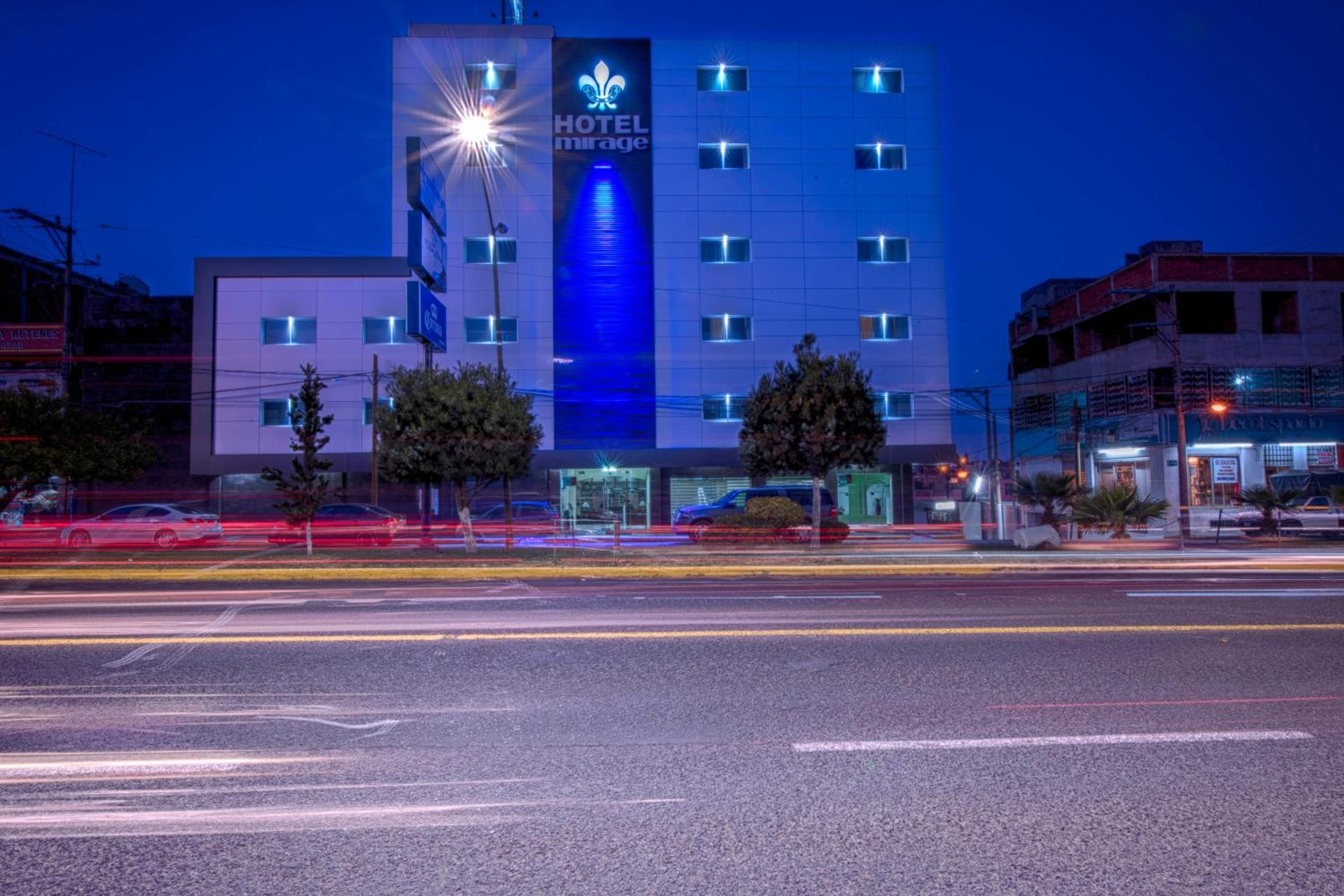 Ein mehrstöckiges Hotel mit auffälligem Logo an der Fassade, einem Parkplatz davor und einer Straße mit Lichtspuren, die den Verkehr zeigen.