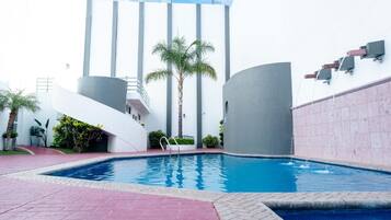 Ein Pool mit modernem Design, umgeben von einer gepflasterten Fläche mit einer Palme und einem Gebäude im Hintergrund, das eine markante, geschwungene Struktur aufweist.